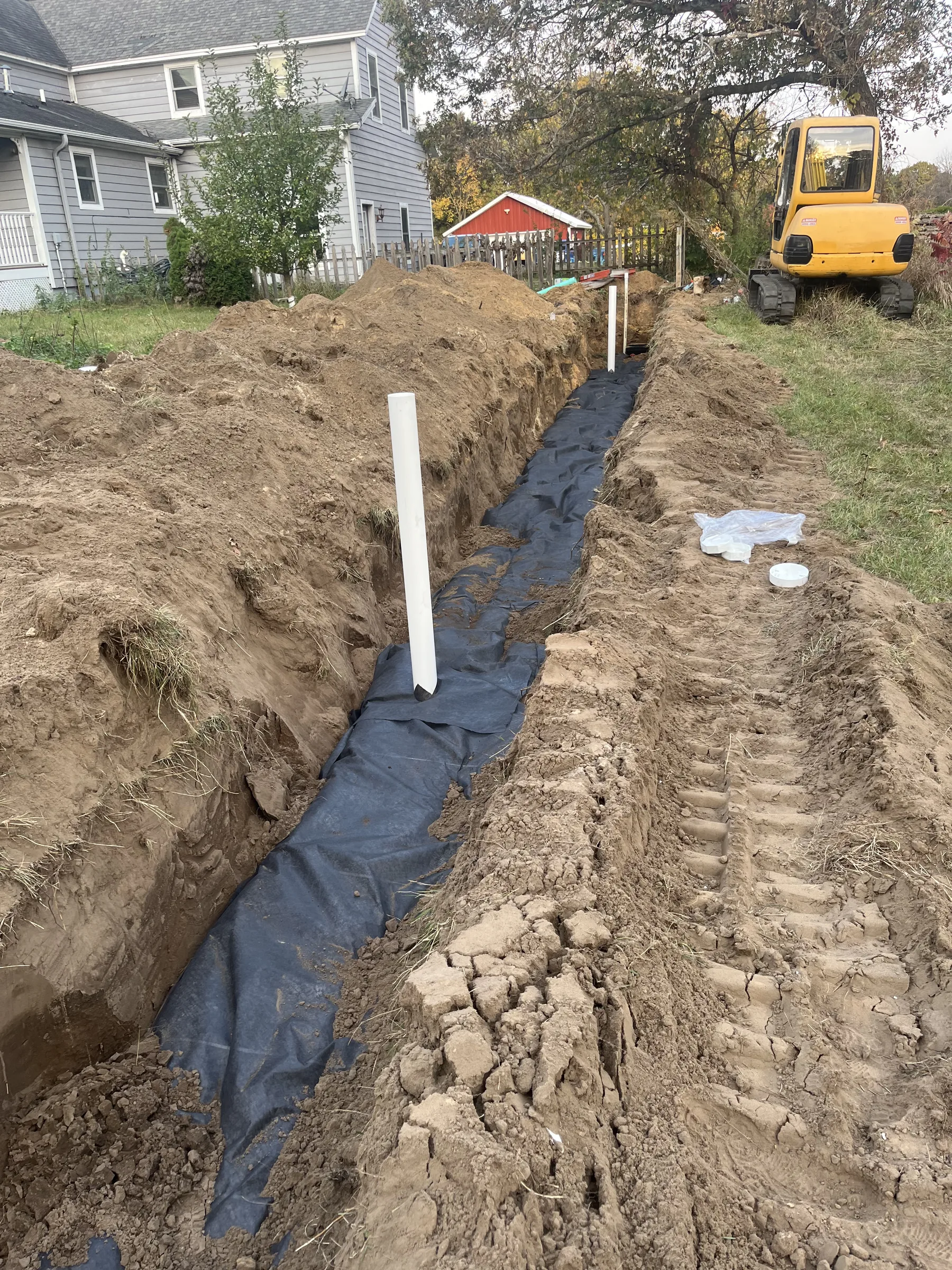 Excavated trench with black drainage liner and white PVC pipe for plumbing installation near residential home.
