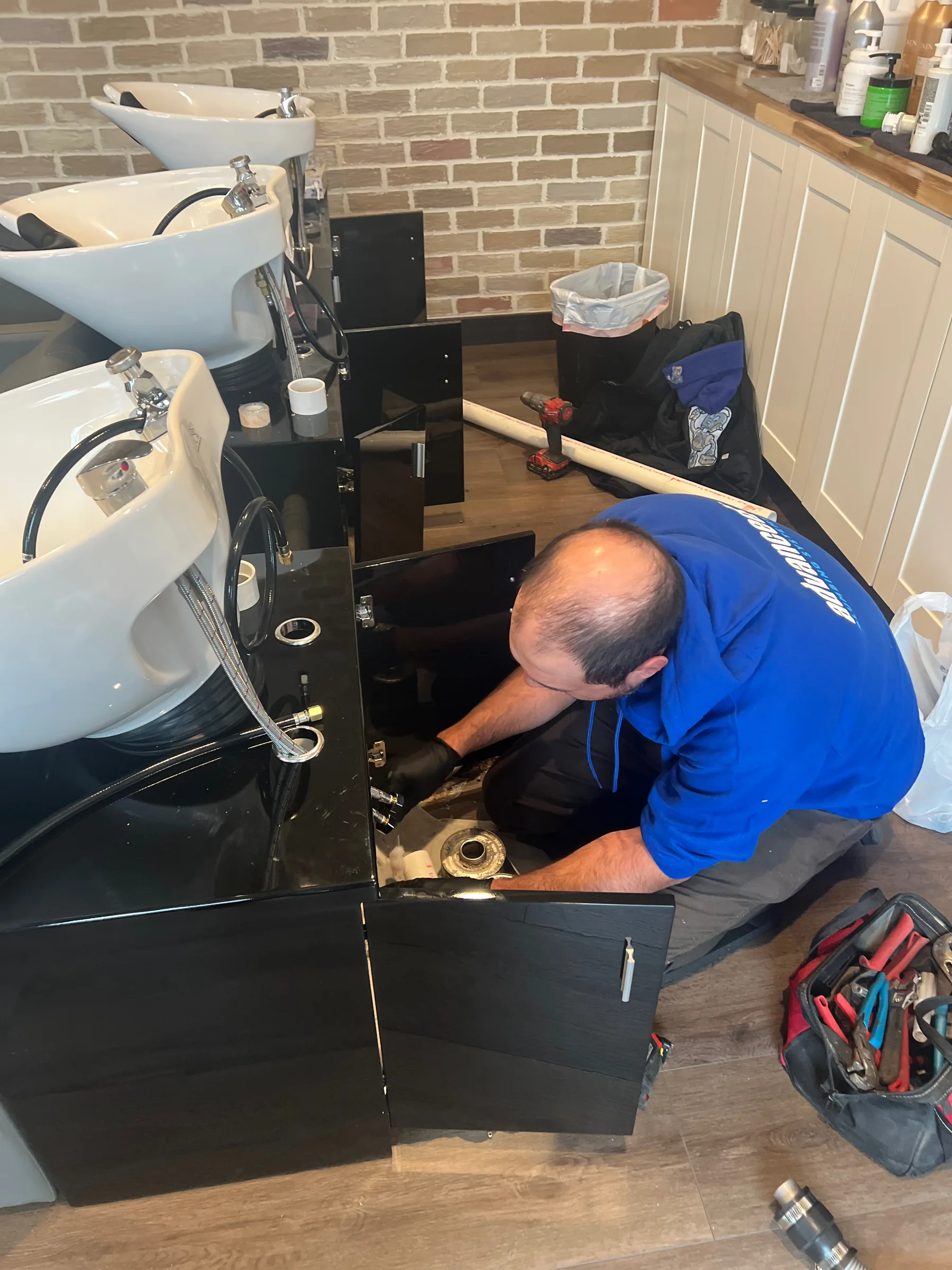Plumber in blue shirt installing or repairing under-sink plumbing in salon bathroom with brick walls.