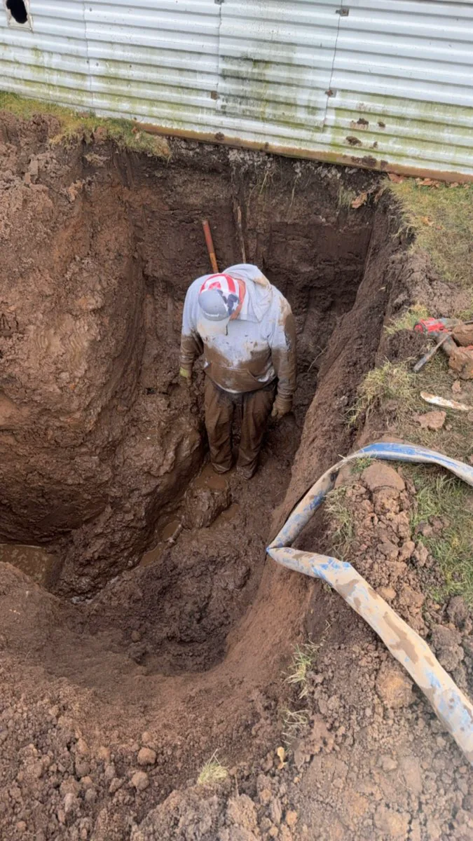 Plumber digging a deep trench in soil next to a metal building foundation.