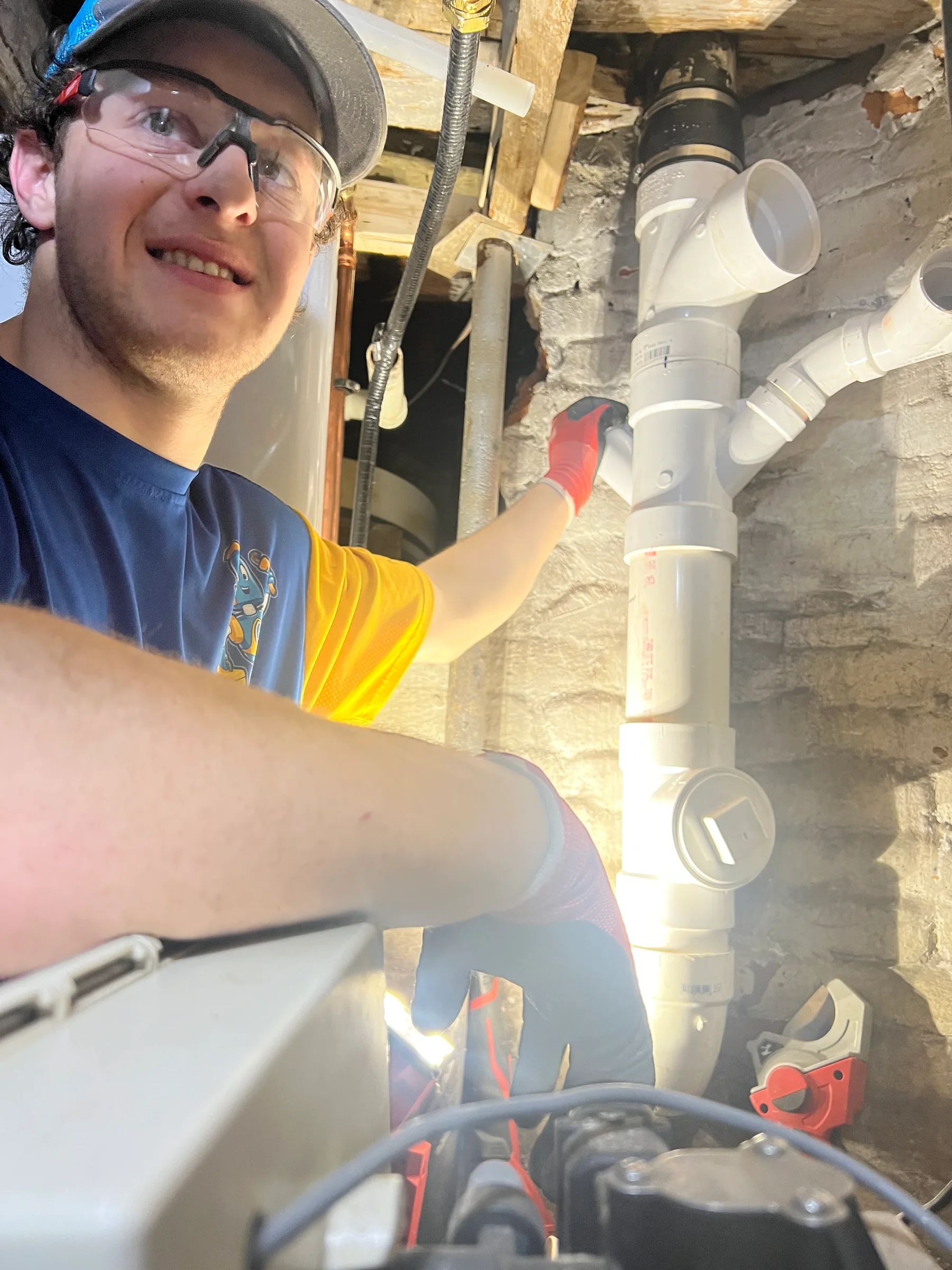Plumber in safety glasses and cap inspecting white PVC pipes in a basement.