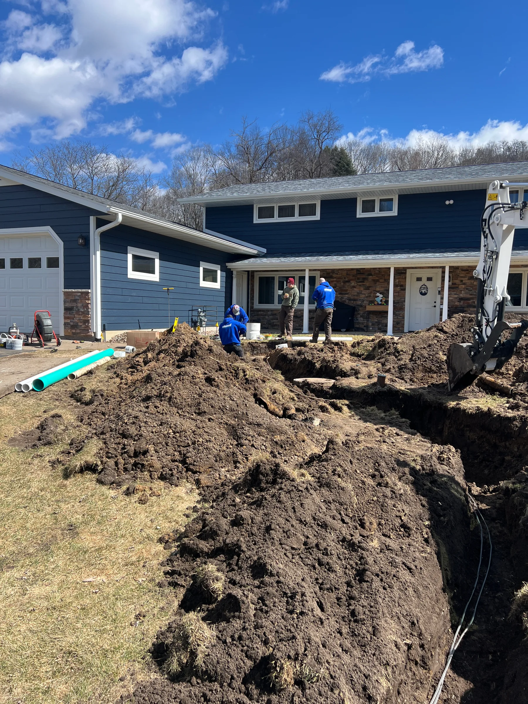 Plumbing crew excavating yard with heavy equipment near blue residential home.