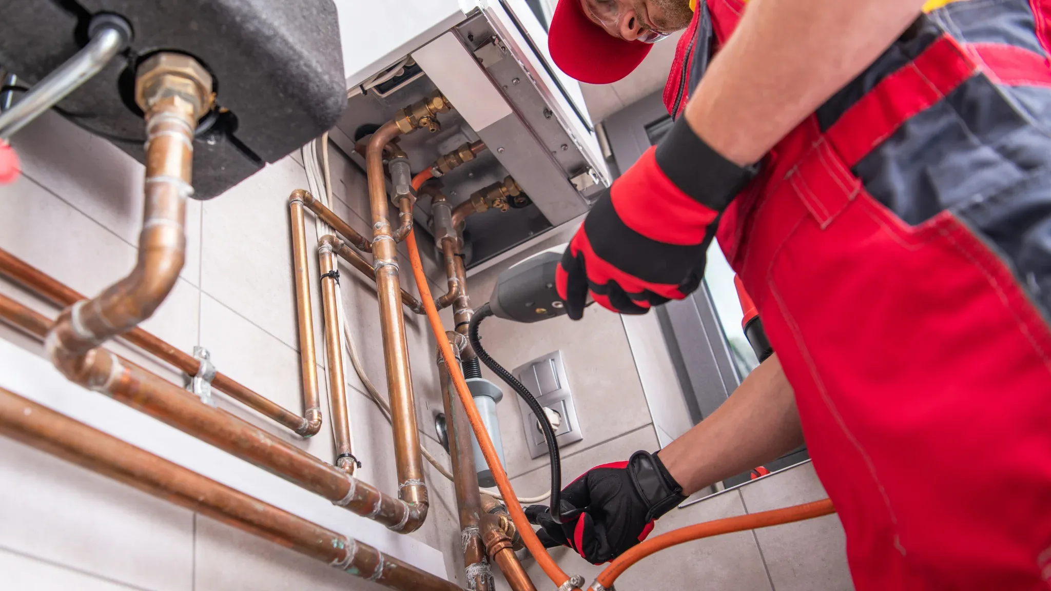 Professional plumber in red work uniform inspecting and servicing a residential water heater system with copper piping connec