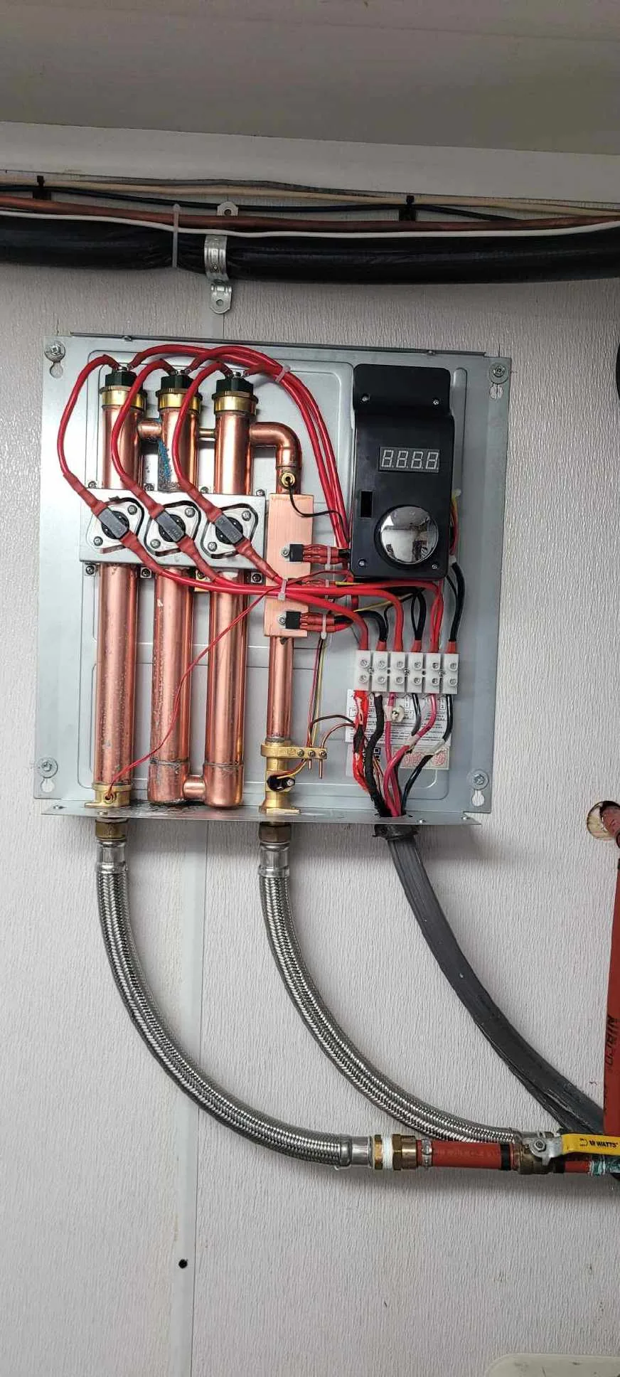 Tankless water heater system with copper pipes, red wiring, and digital control panel mounted on white wall.