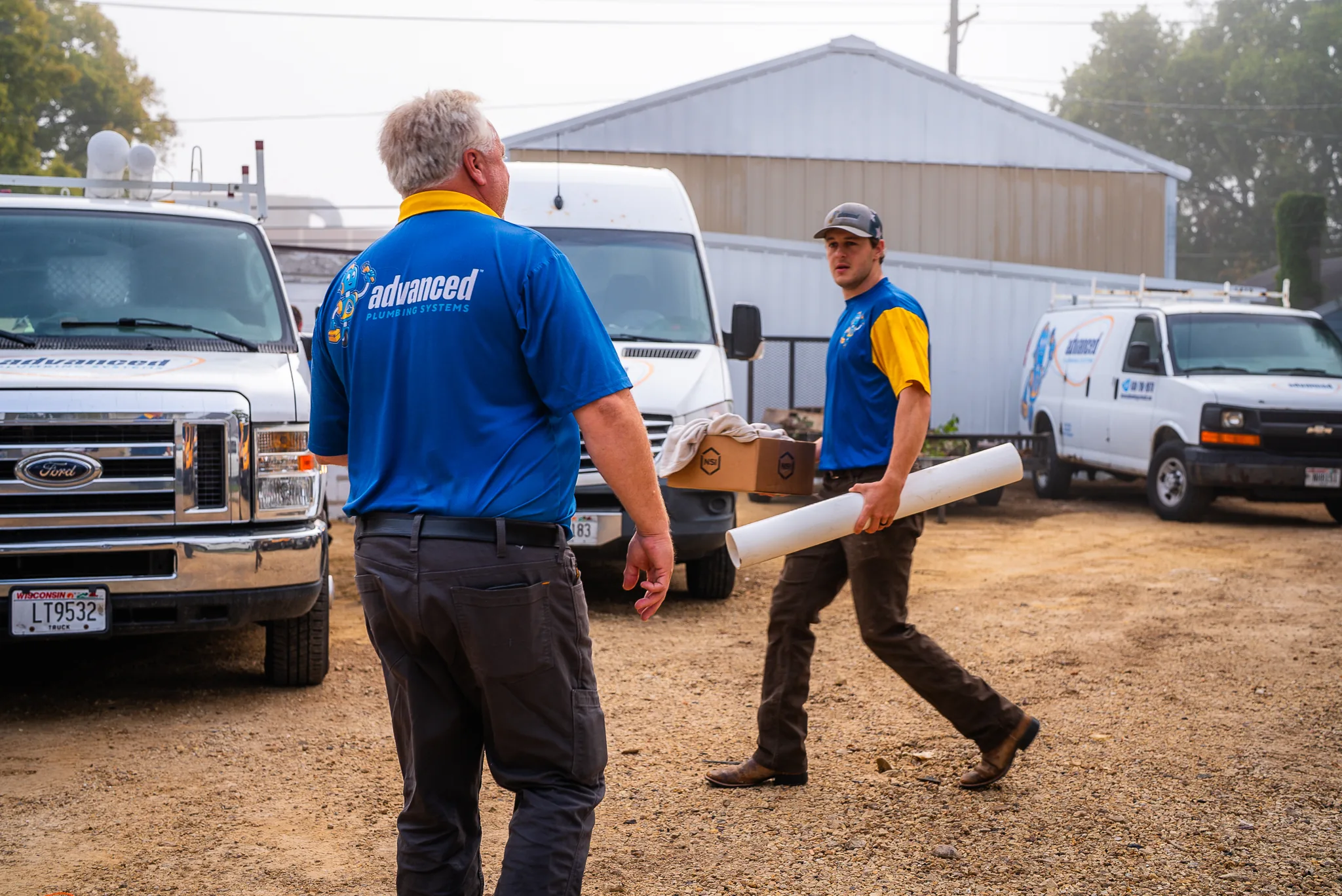 Two plumbers in blue Advanced Plumbing Systems shirts carrying supplies at a job site with service vehicles.