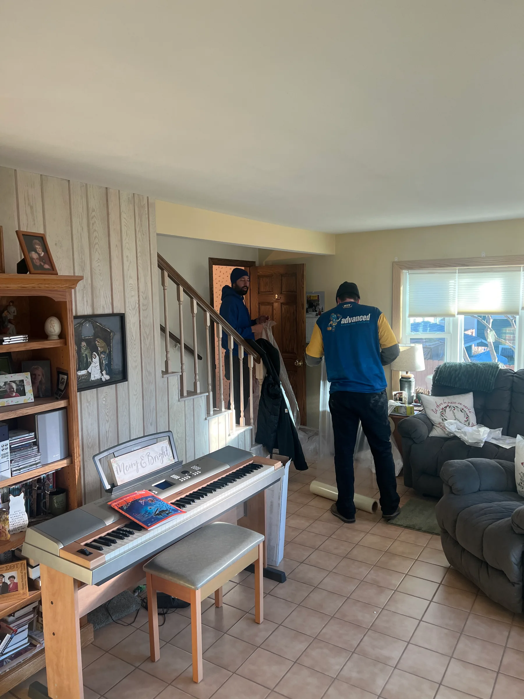 Two plumbers in blue and yellow uniforms working inside a residential home with a piano and living room furniture.