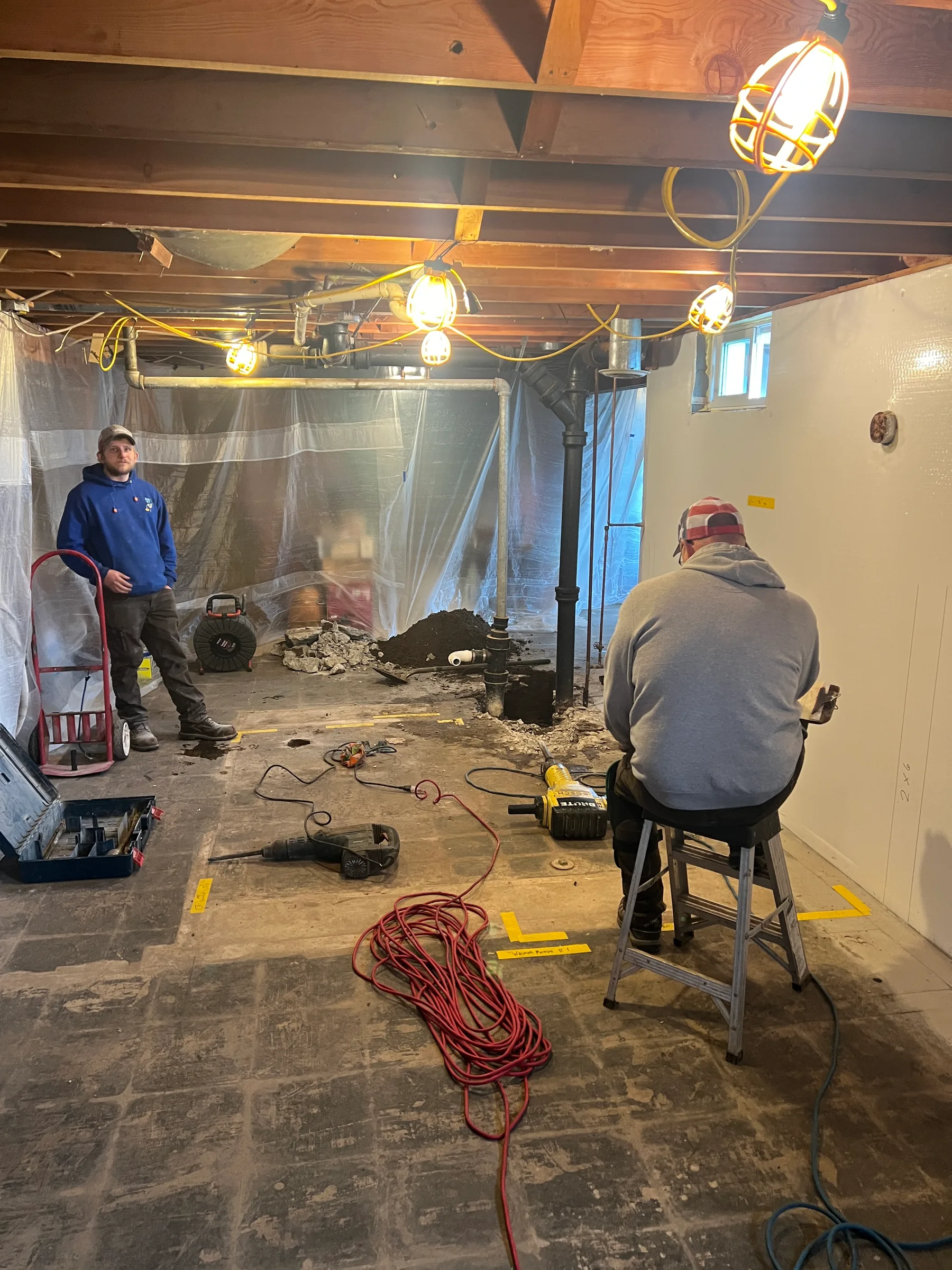 Two plumbers working in an unfinished basement with exposed piping and construction materials.