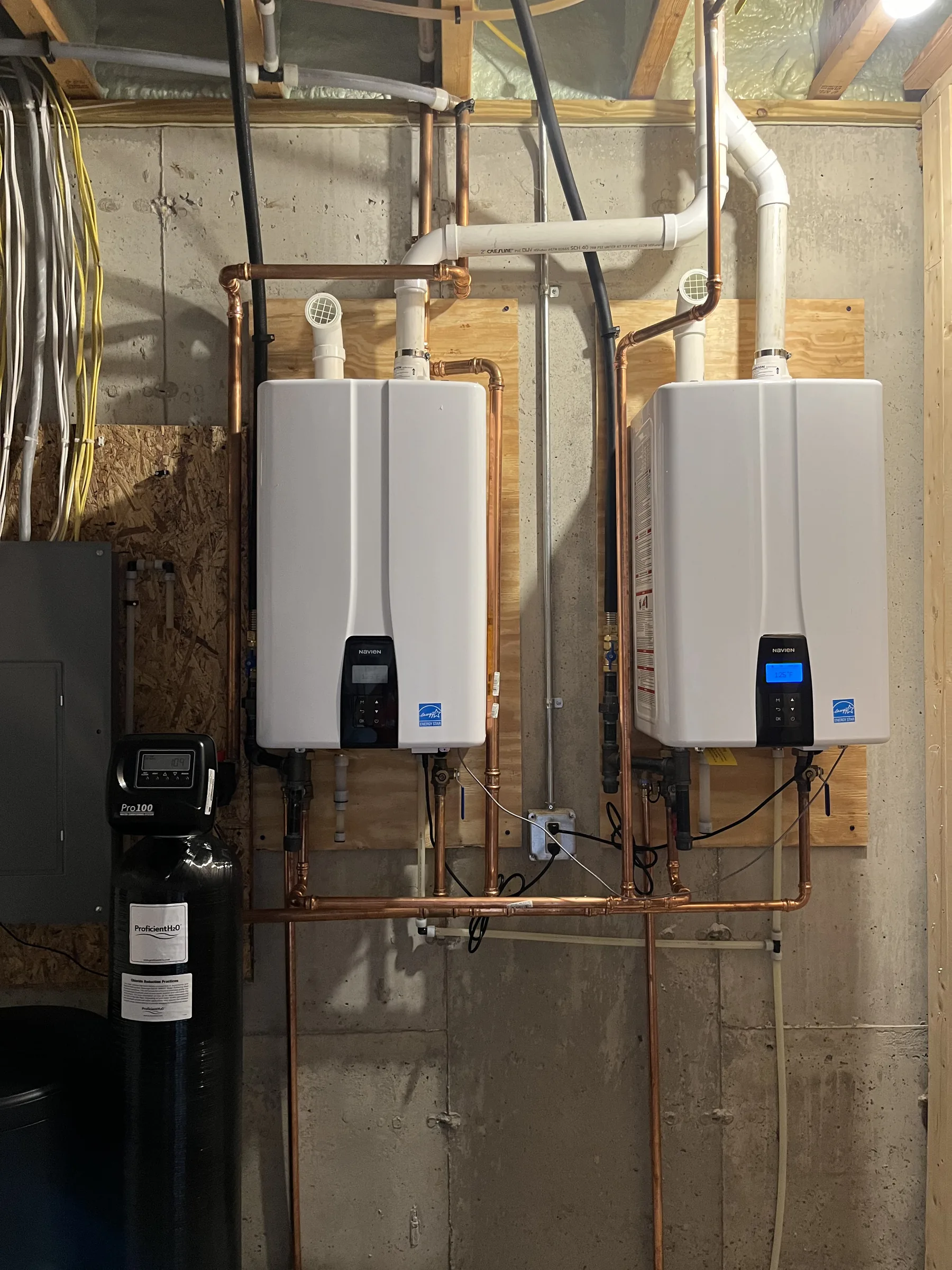 Two white tankless water heaters with digital displays mounted on a basement wall with copper piping.